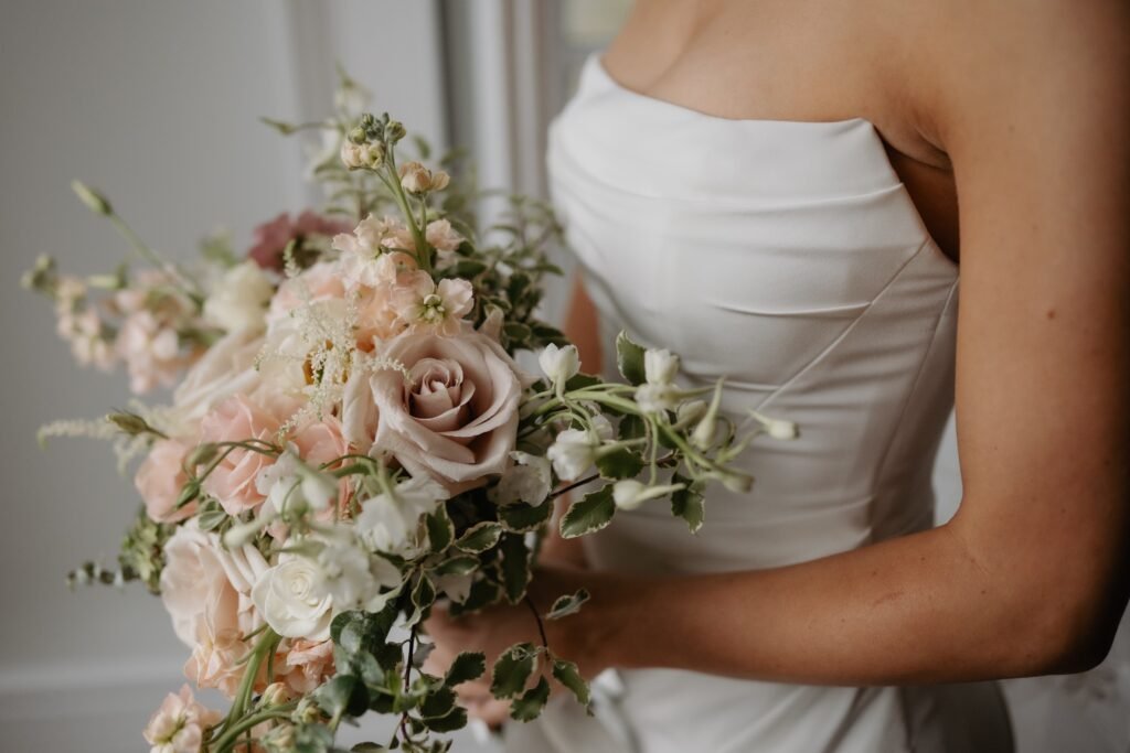 Blush bridal bouquet with mauve roses, stock, and variegated greenery held by a bride in a strapless gown